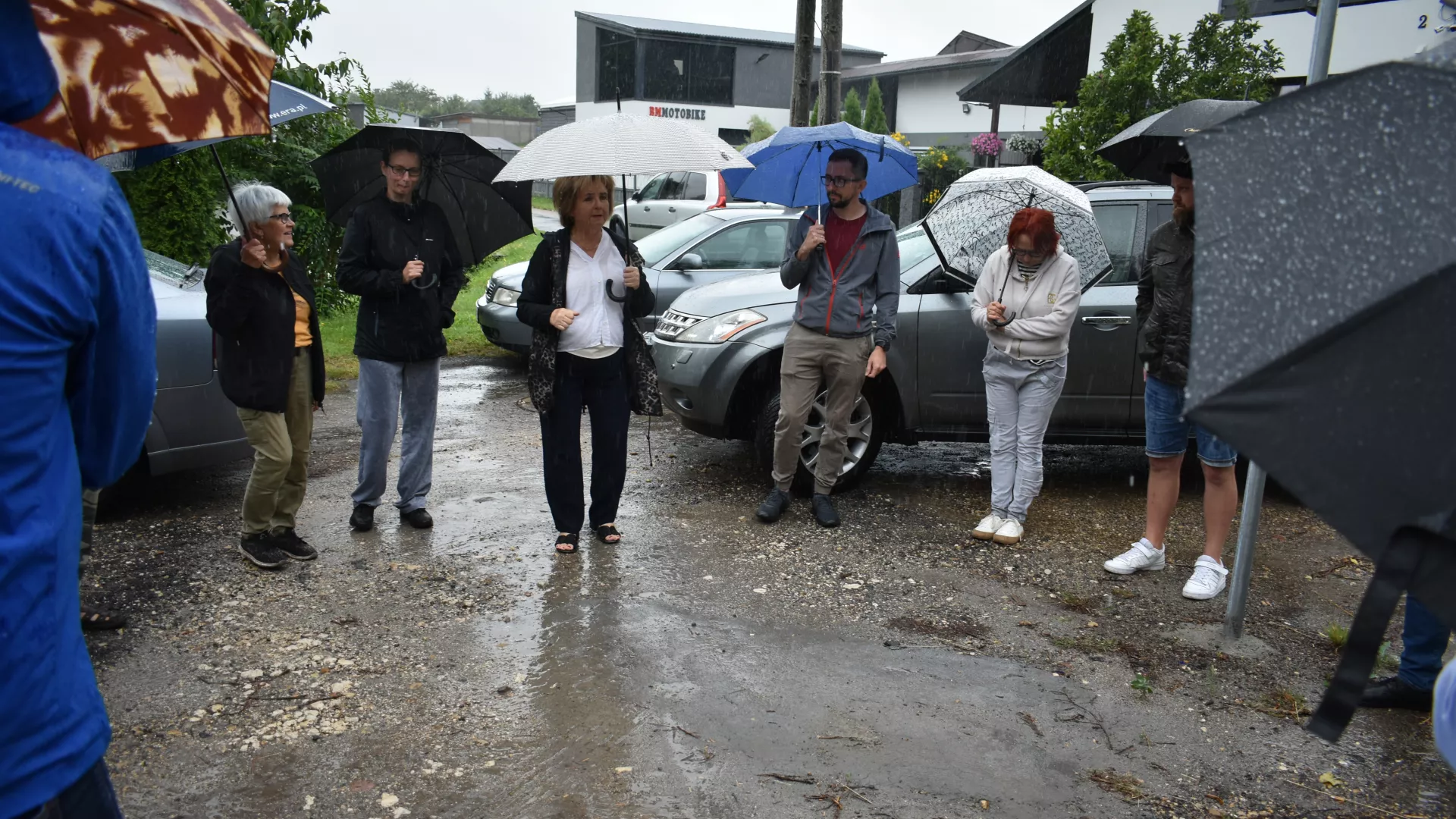 Spotkanie z przedstawicielami GDDKiA w Parzymiechach przy skrzyżowaniu drogi krajowej z ulicą Starowiejską. Na zdjęciu m.in. Wójt Gminy Lipie, radni z Parzymiech i mieszkańcy. Z powodu deszczu większość osób trzyma otwarte parasole. W tle widoczne zaparkowane samochody.