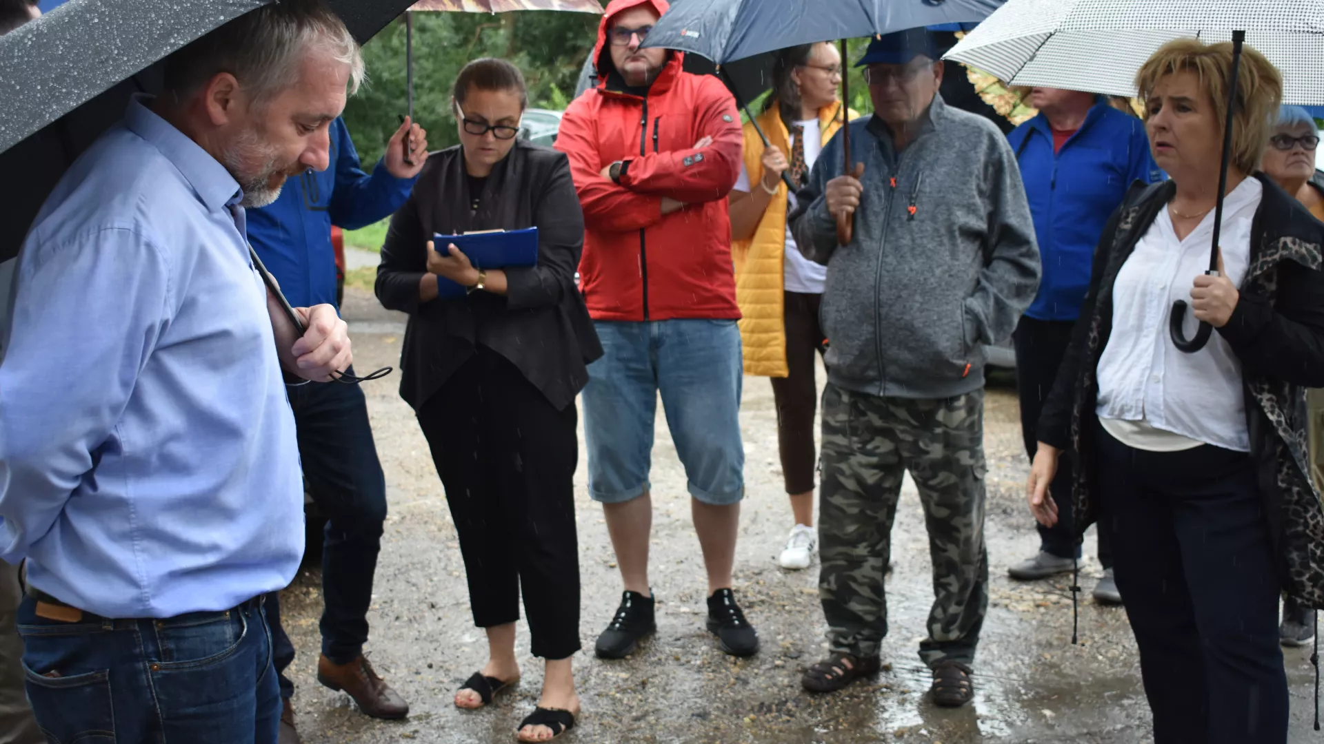 Spotkanie z przedstawicielami GDDKiA w Parzymiechach. Wójt Gminy Lipie rozmawia z przedstawicielem GDDKiA z Katowic. Na drugim planie inni uczestnicy spotkania. Część osób trzyma w ręku otwarte parasole.
