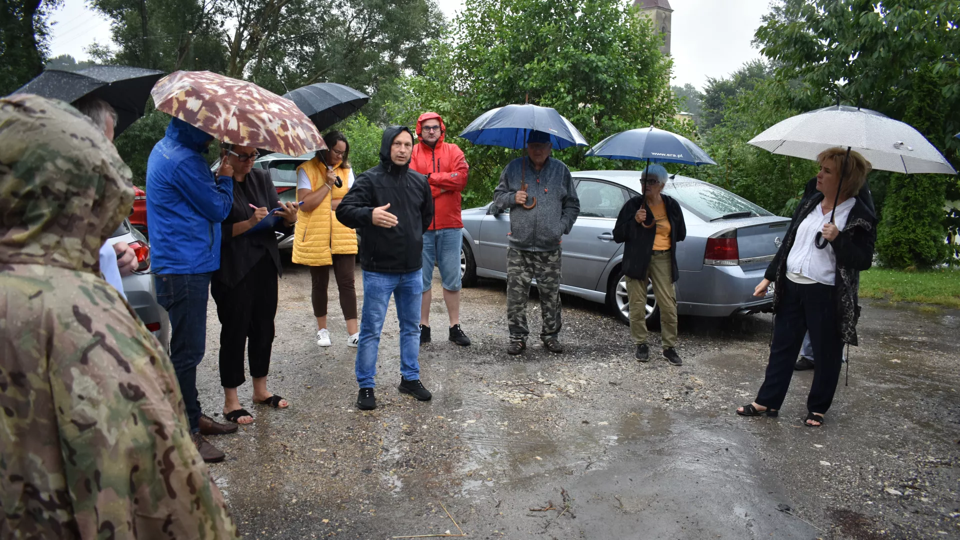 Spotkanie z przedstawicielami GDDKiA w Parzymiechach. Na zdjęciu m.in. Wójt Gminy Lipie, sołtys sołectwa Parzymiechy, pracownicy urzędu gminy i mieszkańcy. Głos zabiera sołtys Parzymiech. Część osób trzyma otwarte parasole. W tle widoczne drzewa i dzwonnica kościoła w Parzymiechach.