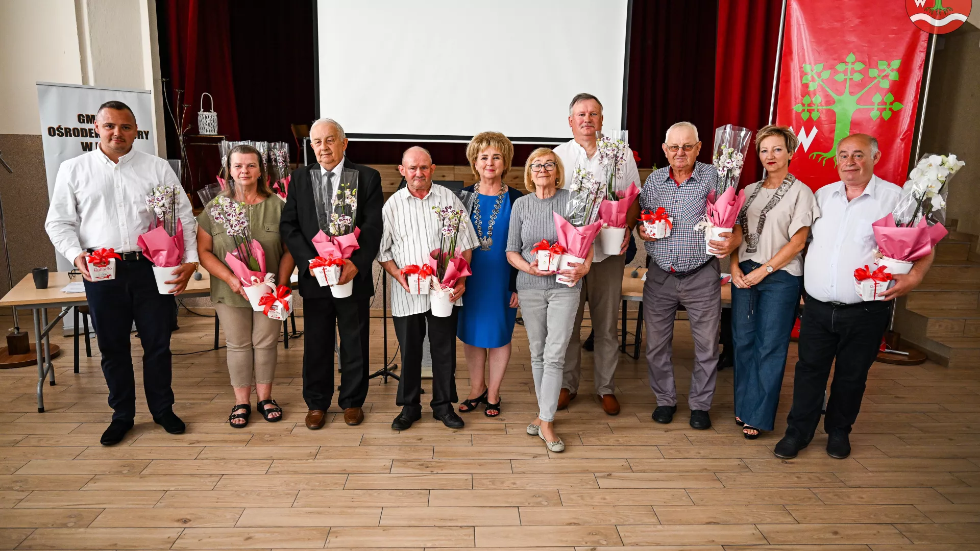 Na zdjęciu znajduje się grupa osób, w tym ustępujący radni i sołtysi, stojący na tle sali z czerwonymi zasłonami i ekranem projekcyjnym. W środku grupy stoi wójt gminy Lipie w niebieskiej sukience. Wszyscy trzymają doniczki z kwiatami, zapakowane w różowy papier. W tle widoczny jest czerwony baner z herbem gminy. Wszyscy uśmiechają się do kamery, tworząc uroczystą atmosferę