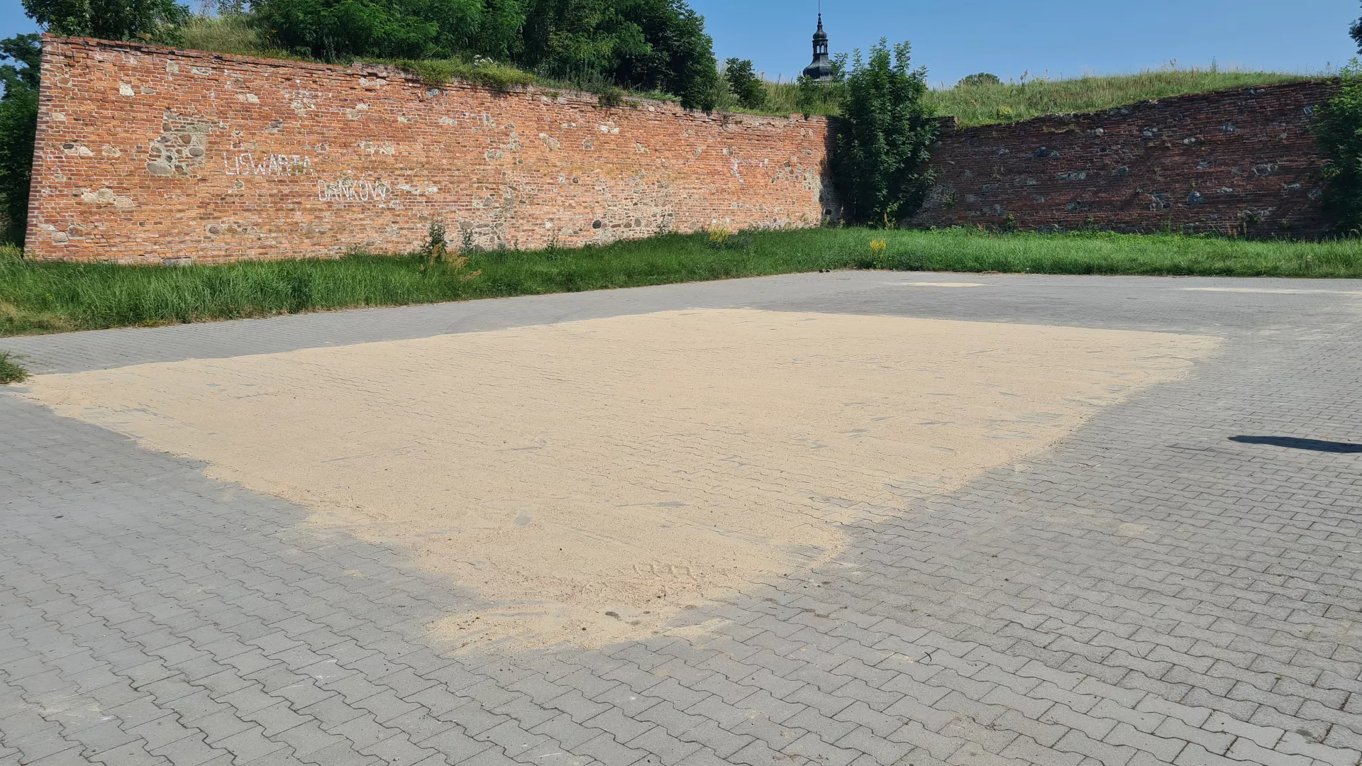 Obszerny plac w sąsiedztwie murów obronnych zamku w Dankowie, utwardzony kostką brukową. Widoczny piasek pokrywający fragmenty powierzchni poddane renowacji.