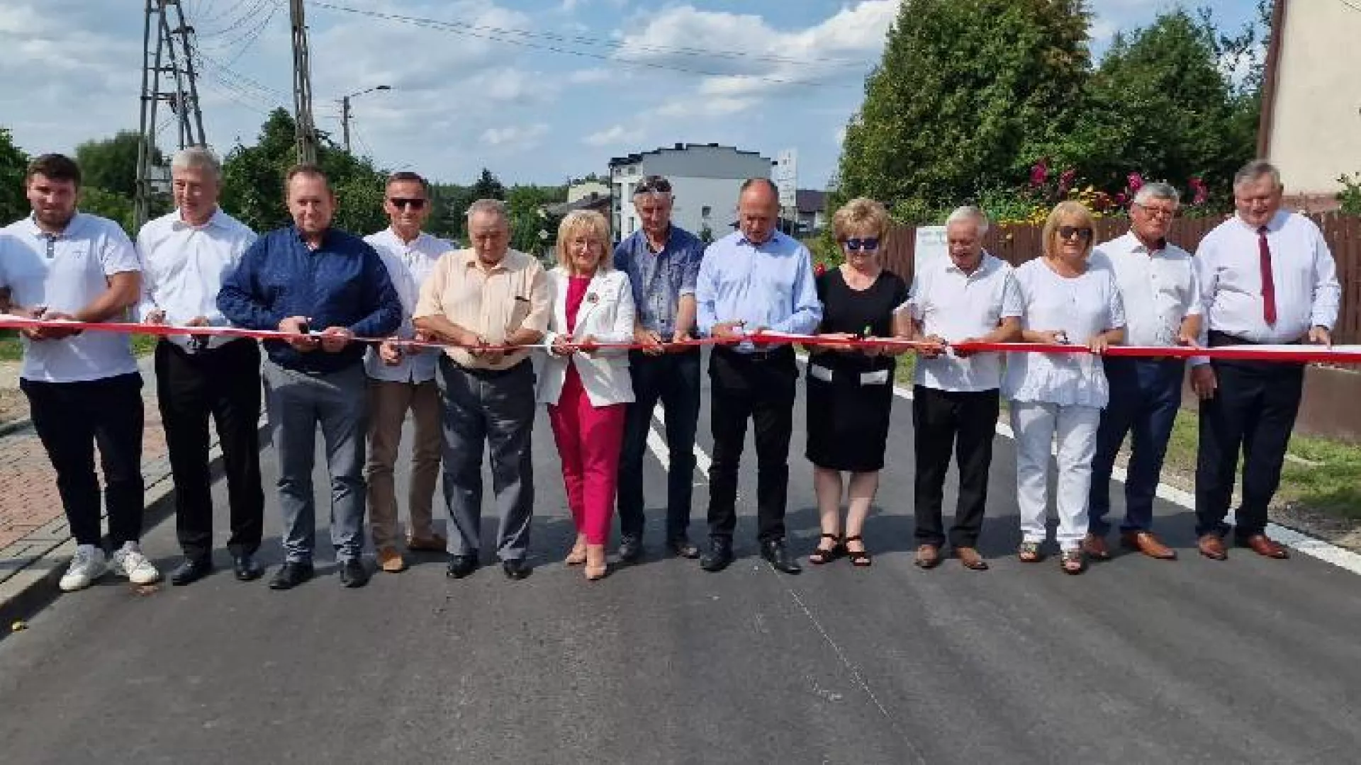 Uczestnicy ceremonii otwarcia drogi powiatowej przygotowują się do przecięcia biało-czerwonej wstęgi rozpostartej w poprzek nowej jezdni.