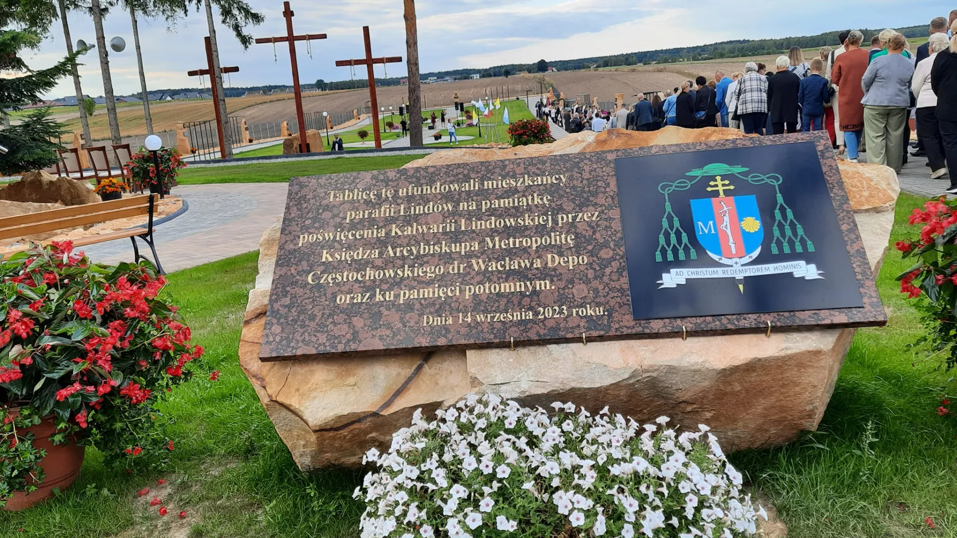 Tablica pamiątkowa z okazji poświęcenia Kalwarii Lindowskiej. W tle – tłum uczestników uroczystości podczas modlitw na terenie kalwarii.