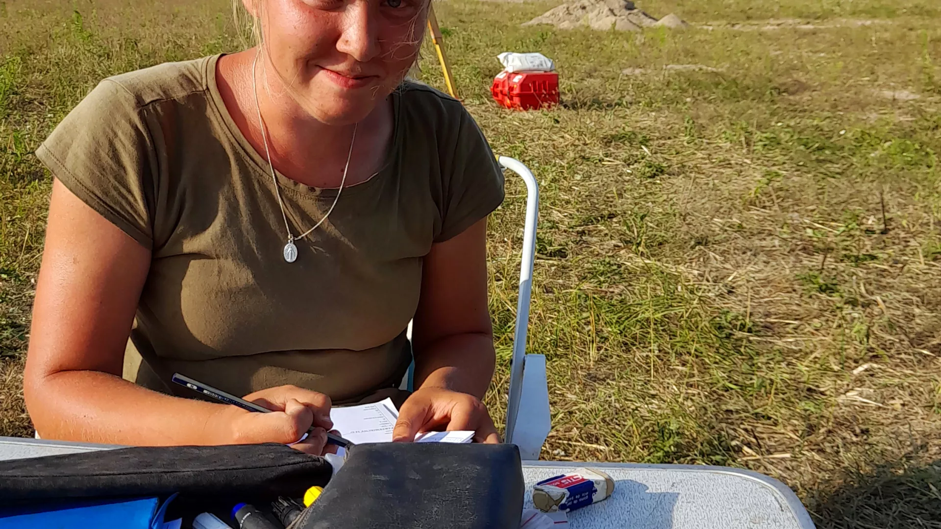 Studentka archeologii Paulina Tarka w warunkach polowych uzupełnia dokumentację badań, siedząc przy rozkładanym stoliku. Każde znalezisko i jego dokładne położenie podlegają skrupulatnemu dokumentowaniu.