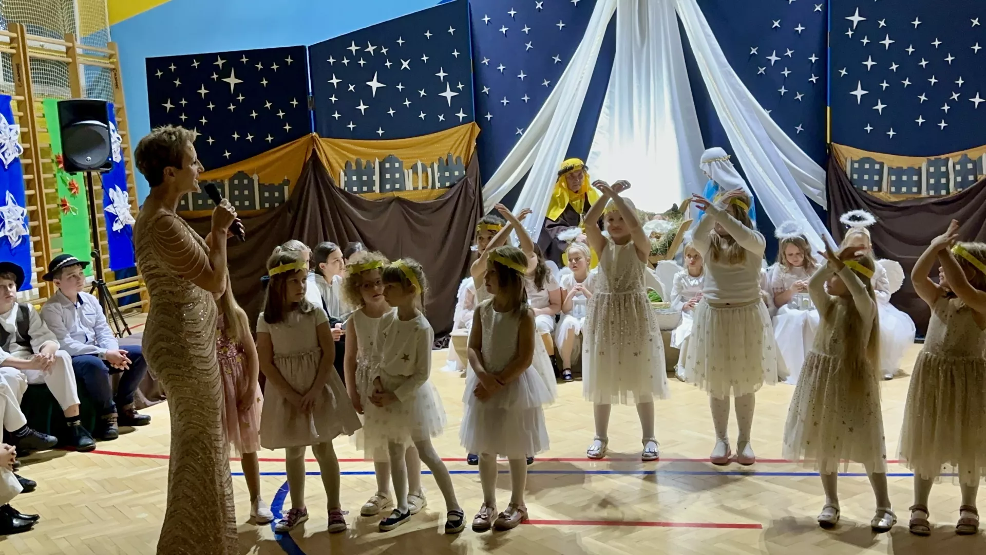 zdjęcie przedstawia aktorów Jasełek ubranych w tradycyjne stroje związane z postaciami bożonarodzeniowymi mi.in. Maryja, św. Józef, aniołowie, pasterze, owieczki,  trzej Królowie, gwiazdki. W tle widać scenografię przedstawienia, która obejmuje miniaturę stajenki betlejemskiej, dekoracje bożonarodzeniowe i jasne oświetlenie tworzące ciepłą i urokliwą atmosferę.  W głównych rolach wystąpili uczniowie wraz z nauczycielami. 