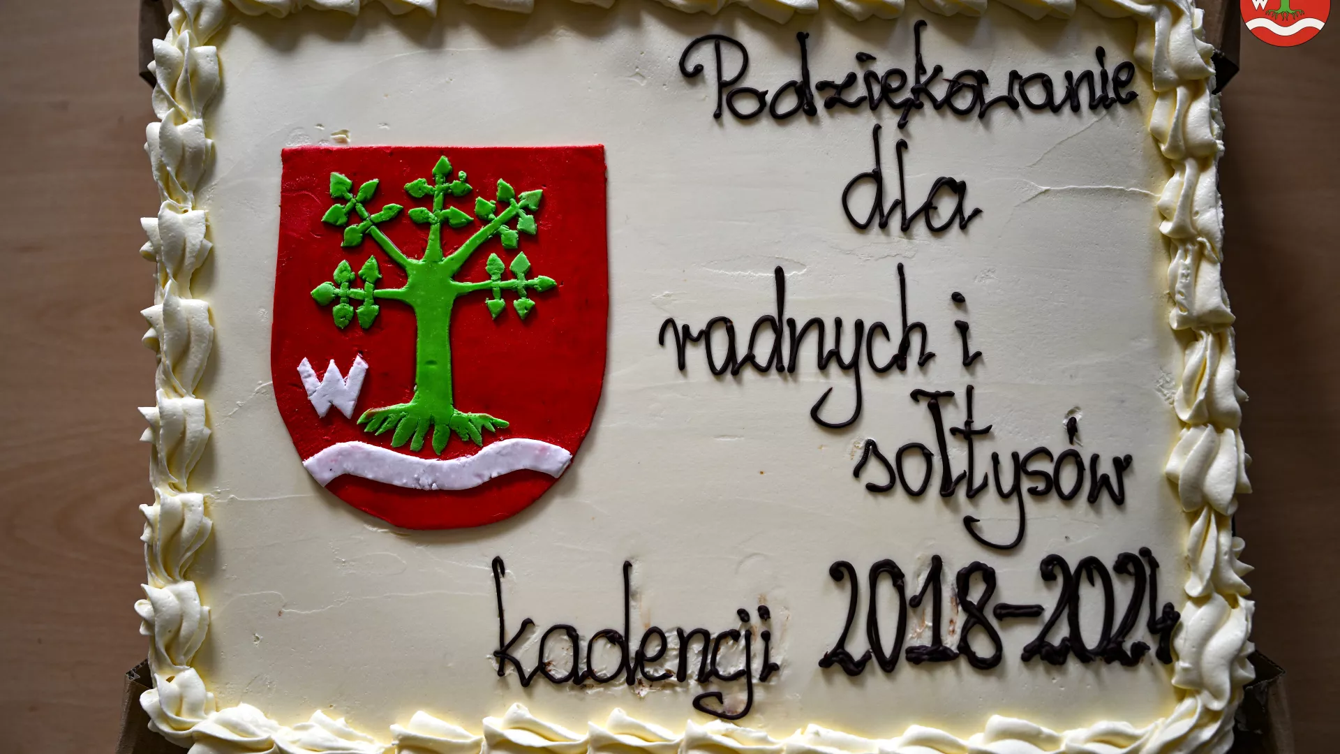 Na zdjęciu widać biały tort ozdobiony kremem, na którym znajduje się herb gminy Lipie oraz napis "Podziękowanie dla radnych i sołtysów kadencji 2018-2024". Tort jest udekorowany eleganckim, kremowym obramowaniem.