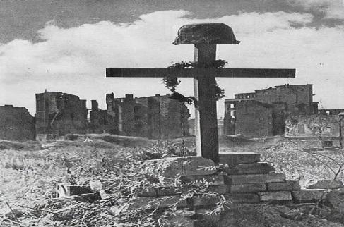 Czarno-białe zdjęcie przedstawia krzyż z wojskowym hełmem na tle ruin zniszczonego miasta.